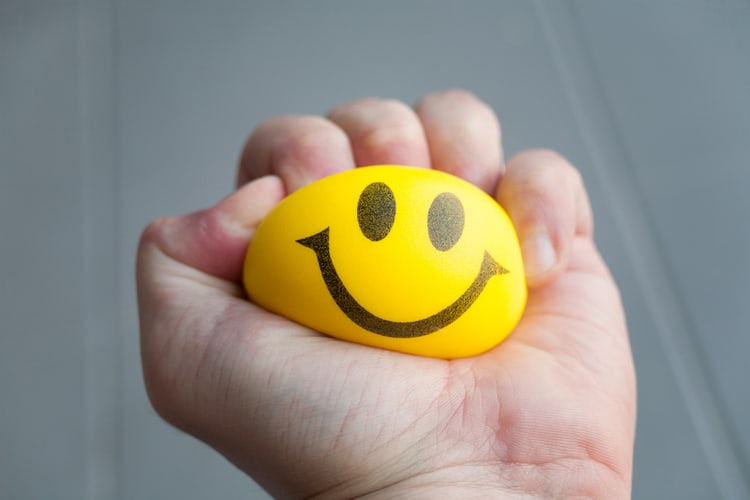 A hand squeezing a yellow stress ball with a smiley face on it