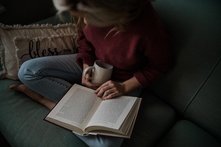A woman sitting on a sofa reading a book with a cup of coffee
