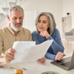 A senior couple sit at a laptop comparing multiple documents