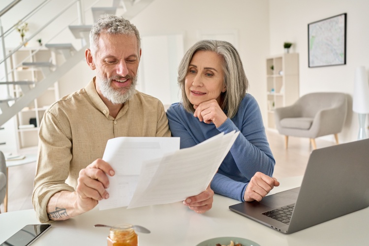 A senior couple sit at a laptop comparing multiple documents